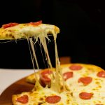 A slice of pepperoni pizza being picked up from the pie, showing off a long cheese pull